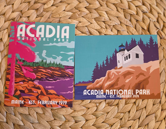Two vintage-style Acadia National Park postcards displayed on a woven round straw mat. One card shows a pink tree and coastal landscape, and the other shows the white Bass Harbor lighthouse house, pine trees, and orange cliffs.