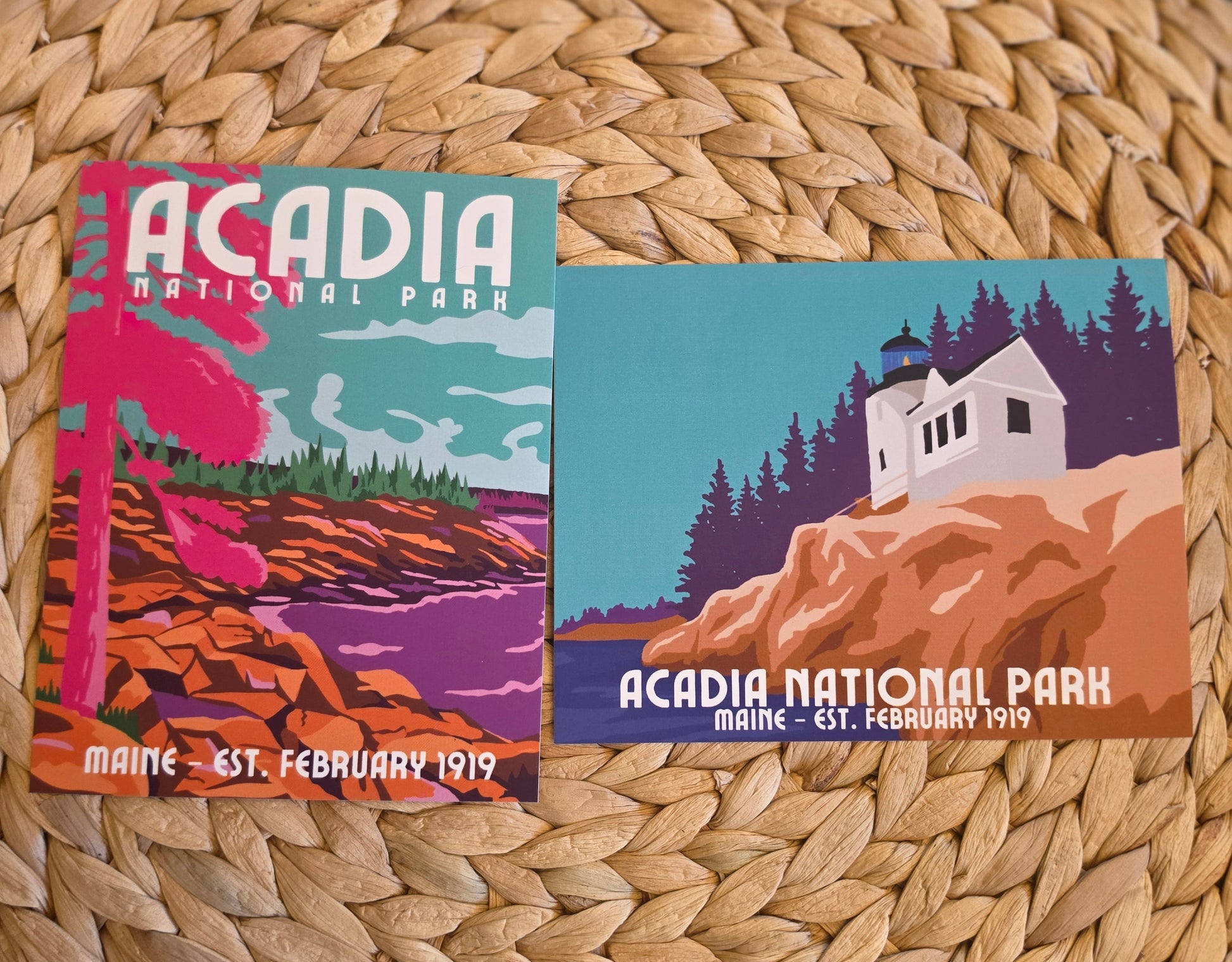 Two vintage-style Acadia National Park postcards displayed on a woven round straw mat. One card shows a pink tree and coastal landscape, and the other shows the white Bass Harbor lighthouse house, pine trees, and orange cliffs.