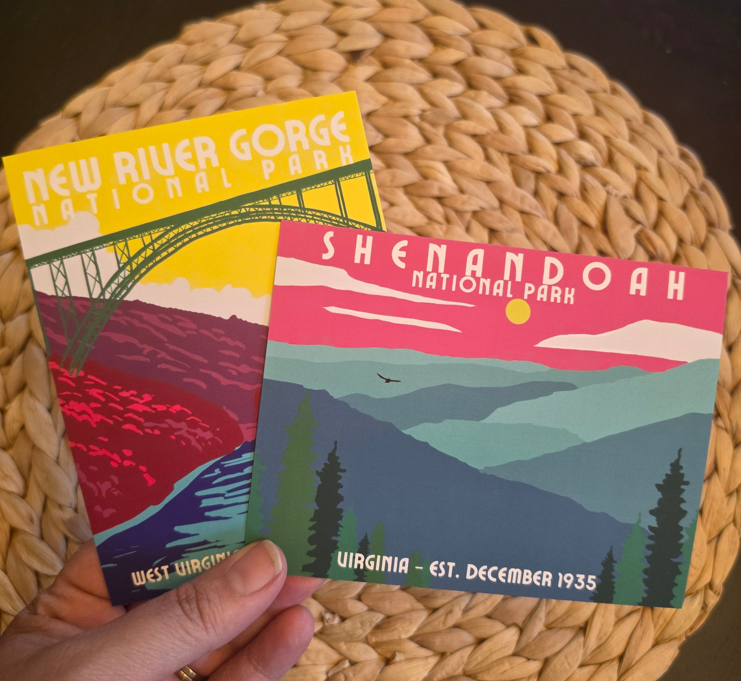 A hand holds two vintage-style National Park postcards over a woven round placemat. One card reads "New River Gorge National Park" and the other reads "Shenandoah National Park" with "Virginia - Est.