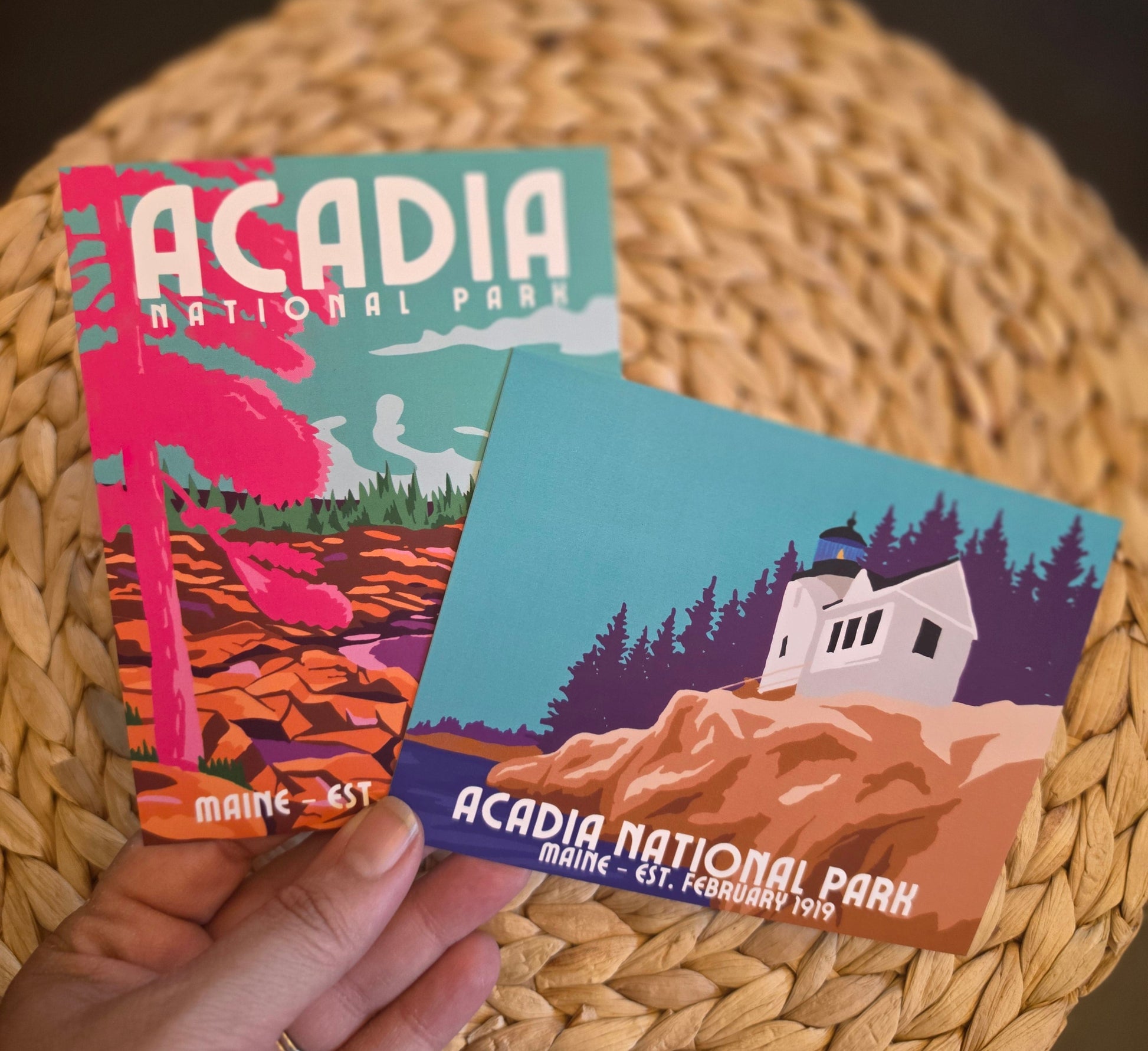 A hand holds two Acadia National Park postcards over a round woven straw placemat on a dark tabletop. The postcards feature colorful vintage-style illustrations of a pink palm tree and a white lighthouse on a rocky cliff surrounded by dark purple pine trees.