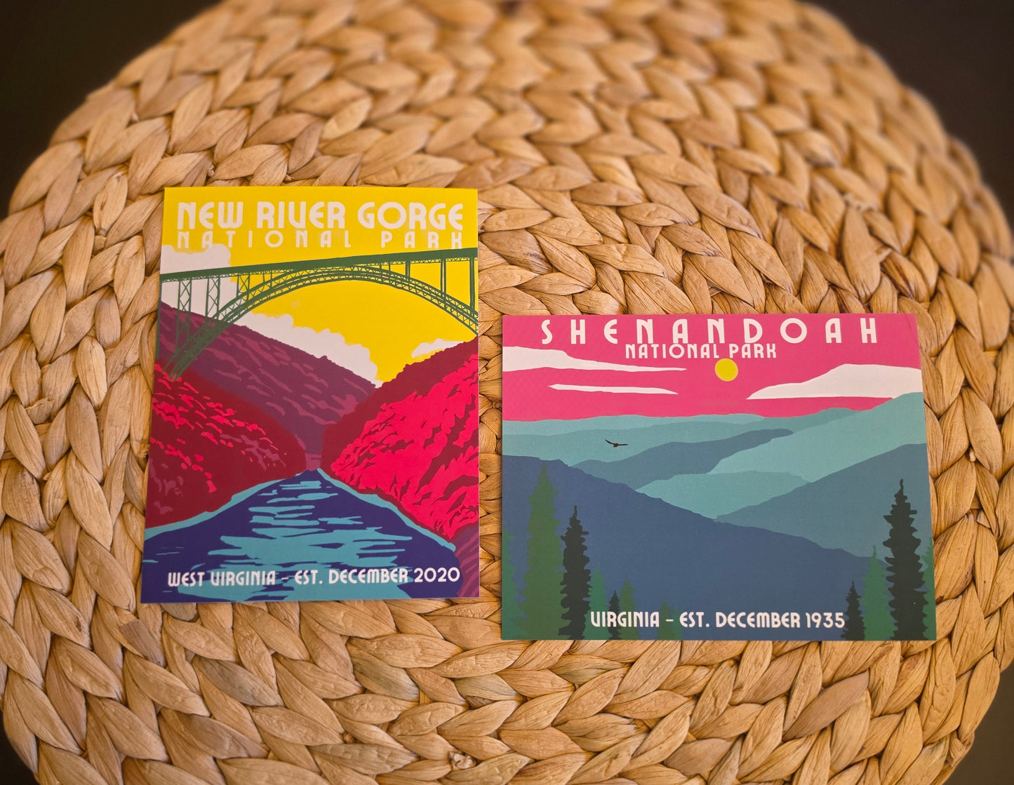 Two colorful National Park postcard-style prints resting on a round woven straw mat against a dark tabletop. The left print reads New River Gorge National Park, and the right print reads Shenandoah National Park.