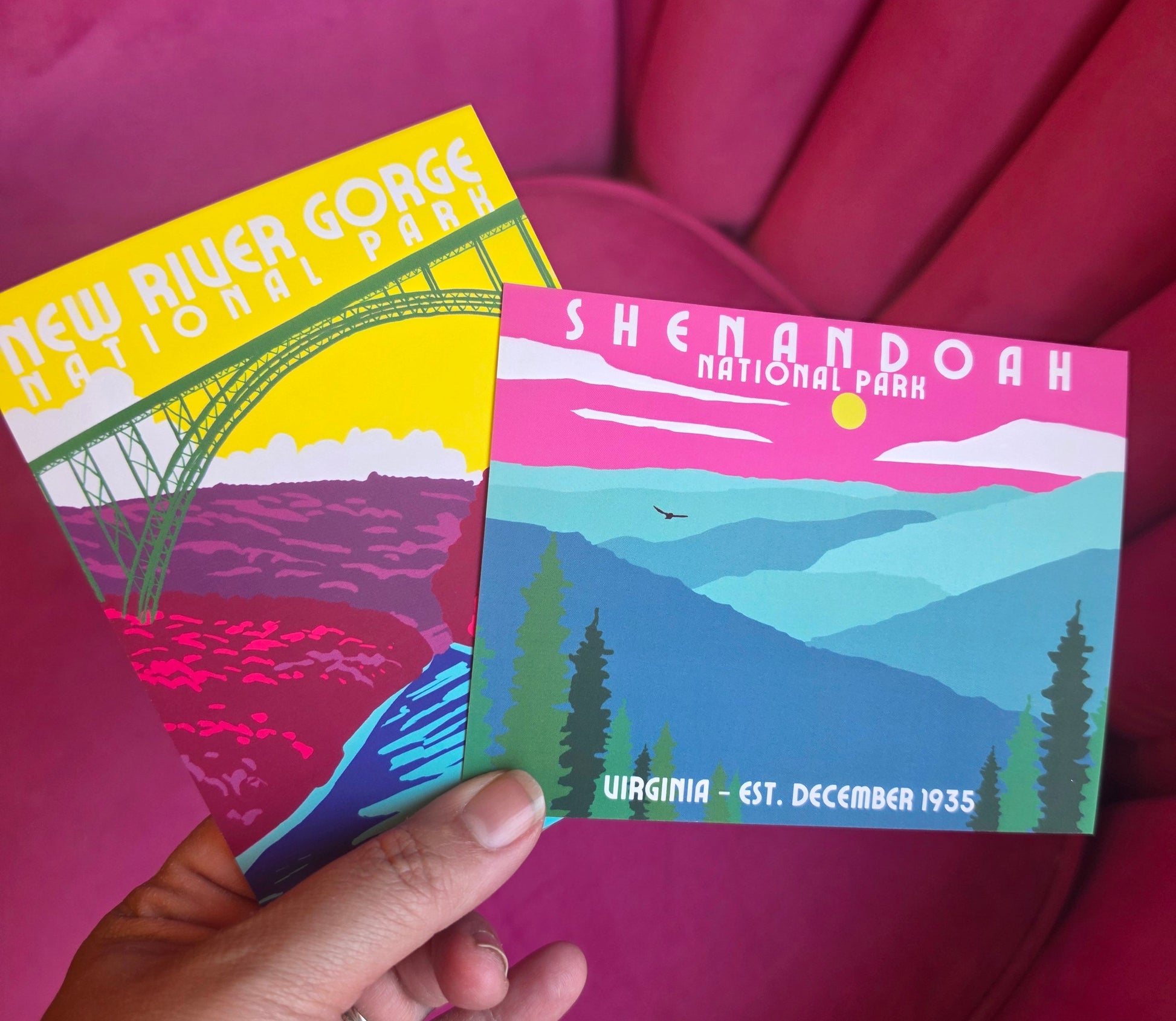 A hand holds two vintage-style National Park postcards in front of a bright pink upholstered chair. One card shows New River Gorge National Park with a yellow bridge and pink landscape, and the other shows Shenandoah National Park with blue mountains and green trees.